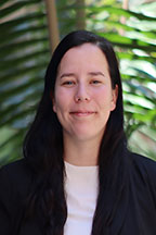 Person with long black hair in a white top and black blazer, standing in front of green foliage.