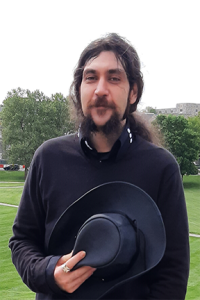 Person holding a black hat, wearing a black sweatshirt, standing outdoors on grass.